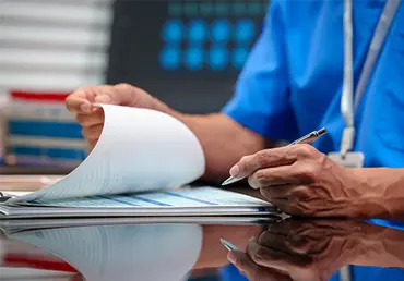 Healthcare worker in blue scrubs reviewing paperwork on clipboard at hospital desk, holding pen, clinical setting with lab supplies and monitor in background.