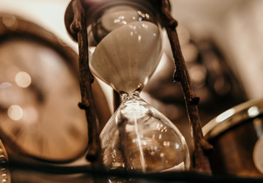 An hour glass is photographed with a clock in the background.