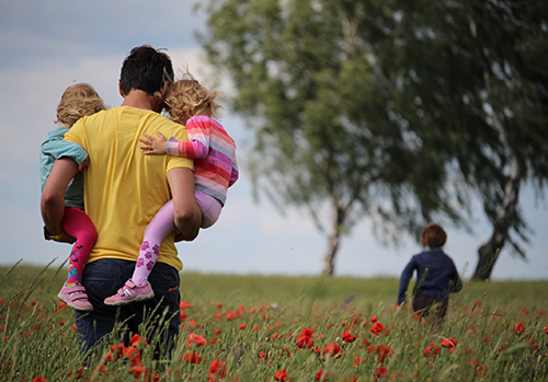 A father carries his two daughters as he walks in a flower field while his son runs.