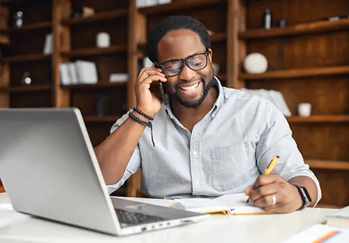 Self-employed man, sitting at his desk working, smiles as he speaks on his cell phone and takes notes. 