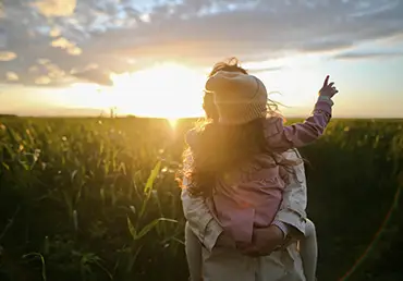 A mom holds her daughter as they enjoy the sunset in a field of tall greenery.