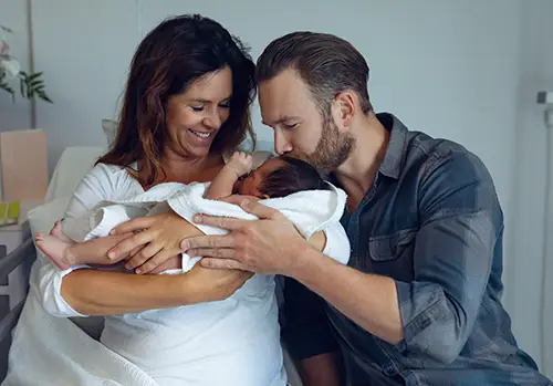 New parents hold brand new baby while the father is kissing the baby on the forehead.