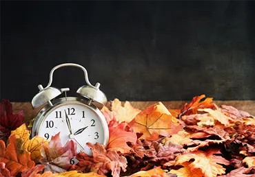 Alarm clock in colorful autumn leaves against a dark background with shallow depth of field. Daylight savings time concept.